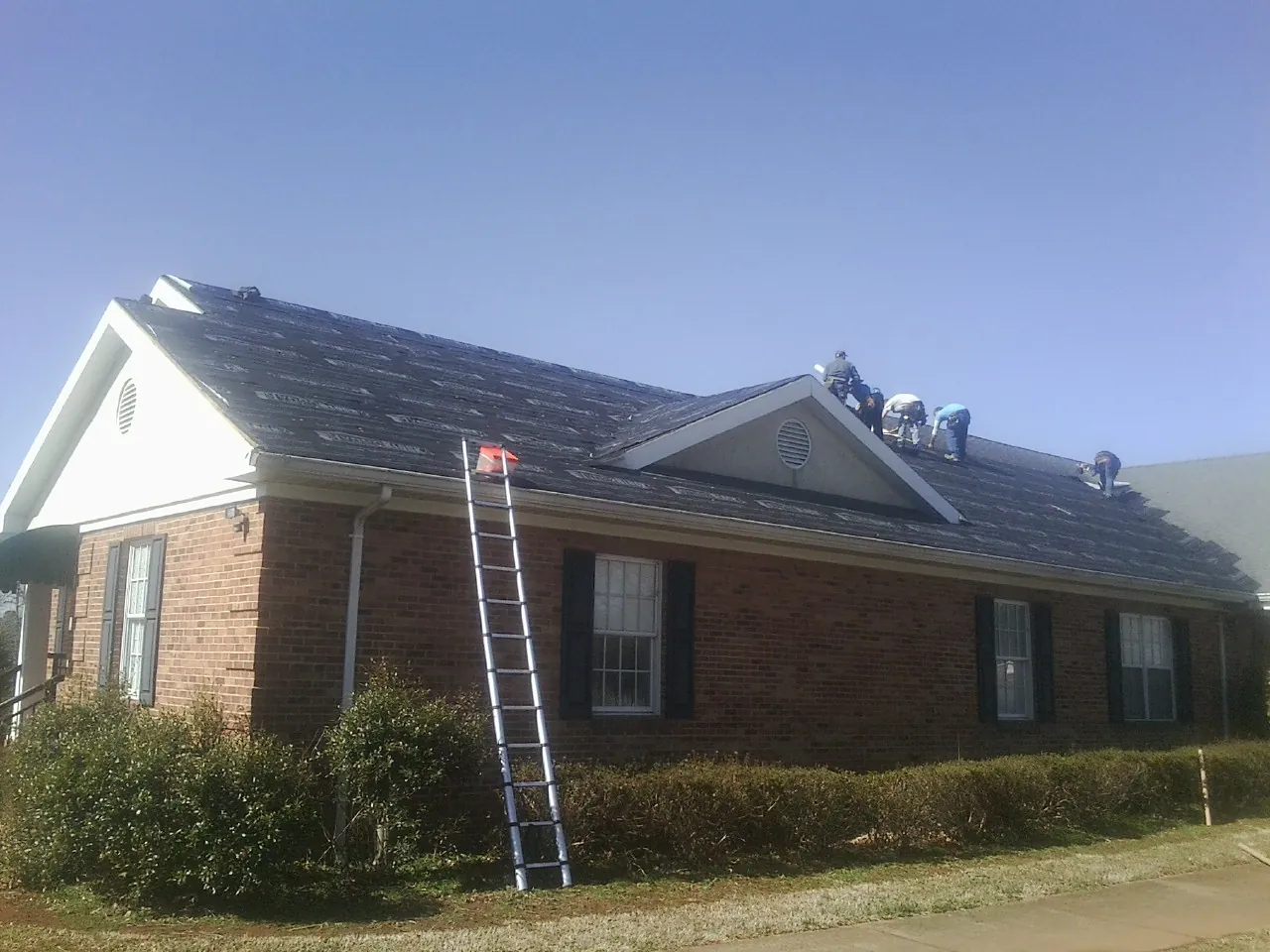 Crew tearing shingles off a brick ranch roof with a ladder set — Emergency Roof Repair work by our crew in Hosford
