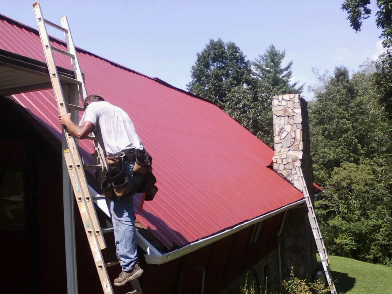 Roofer on a ladder beside a red metal roof and stone chimney — Soffit & Fascia Repair by Roofing Brothers in Hosford